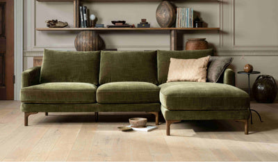 An olive green velvet sectional sofa with a chaise lounge, styled in a warm, modern living room with light wood floors and shelves in the background.