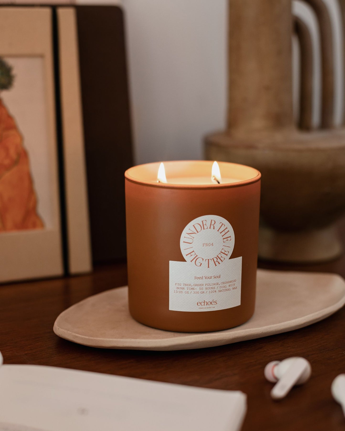 A two-wick candle with a brown label that reads 'Under The Fig Tree' burning inside a clear glass jar on a wooden surface.