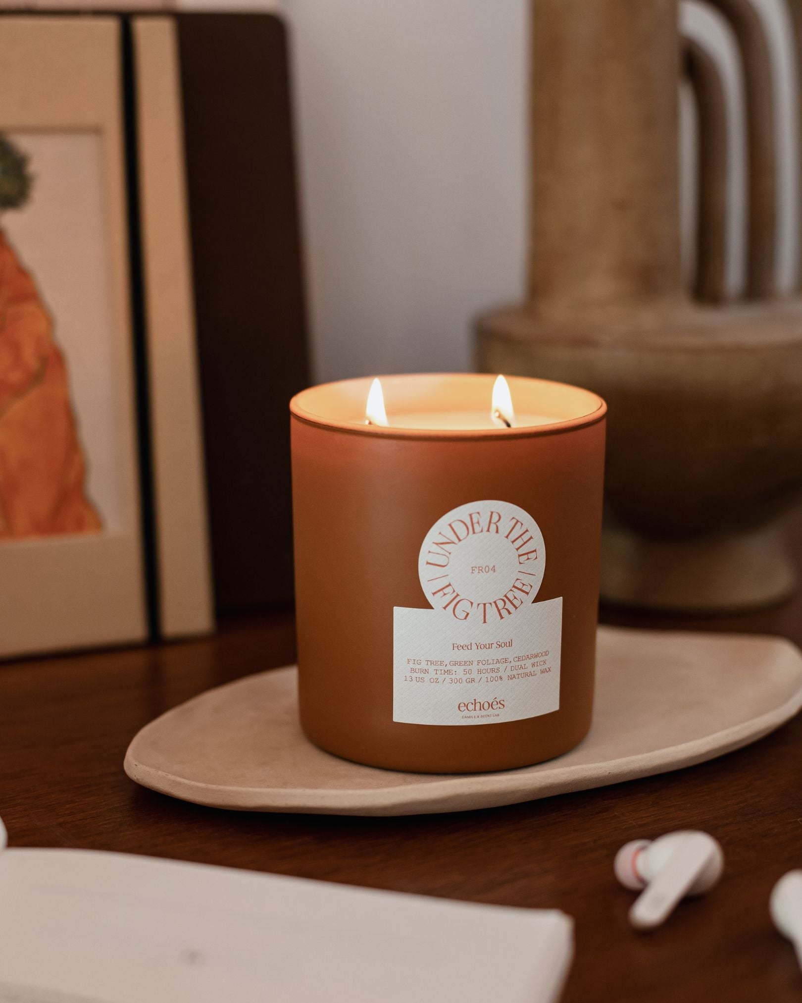 A two-wick candle with a brown label that reads 'Under The Fig Tree' burning inside a clear glass jar on a wooden surface.