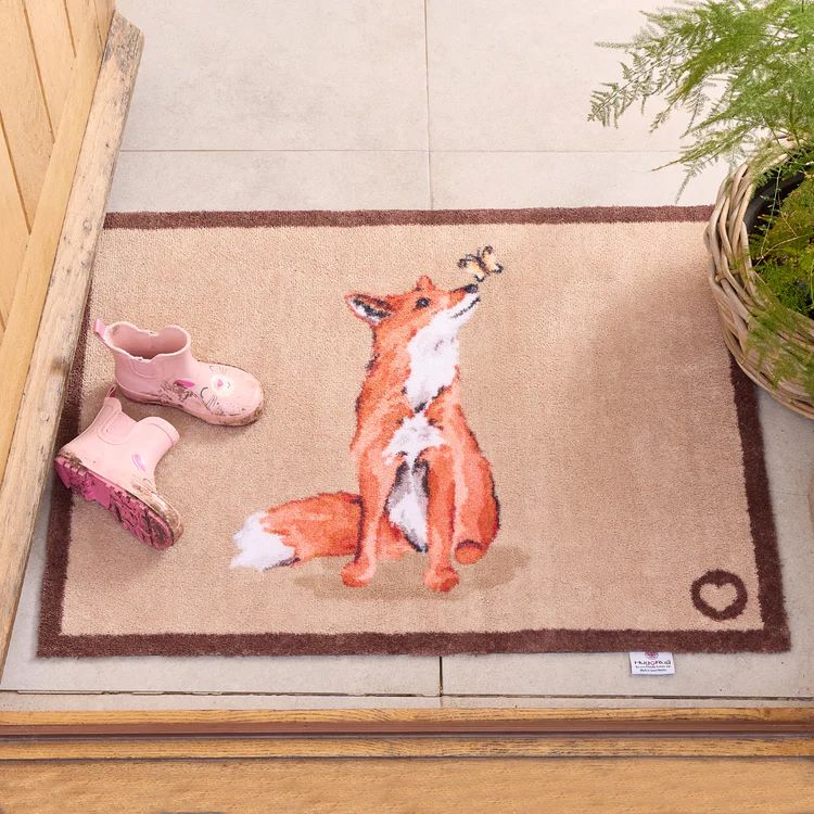 Entry way to a house with a Hug Rug washable mat with a ginger fox design sitting on a neutral background. Kids wellington boots are on the mat and a plantt in a basket is to one side. 