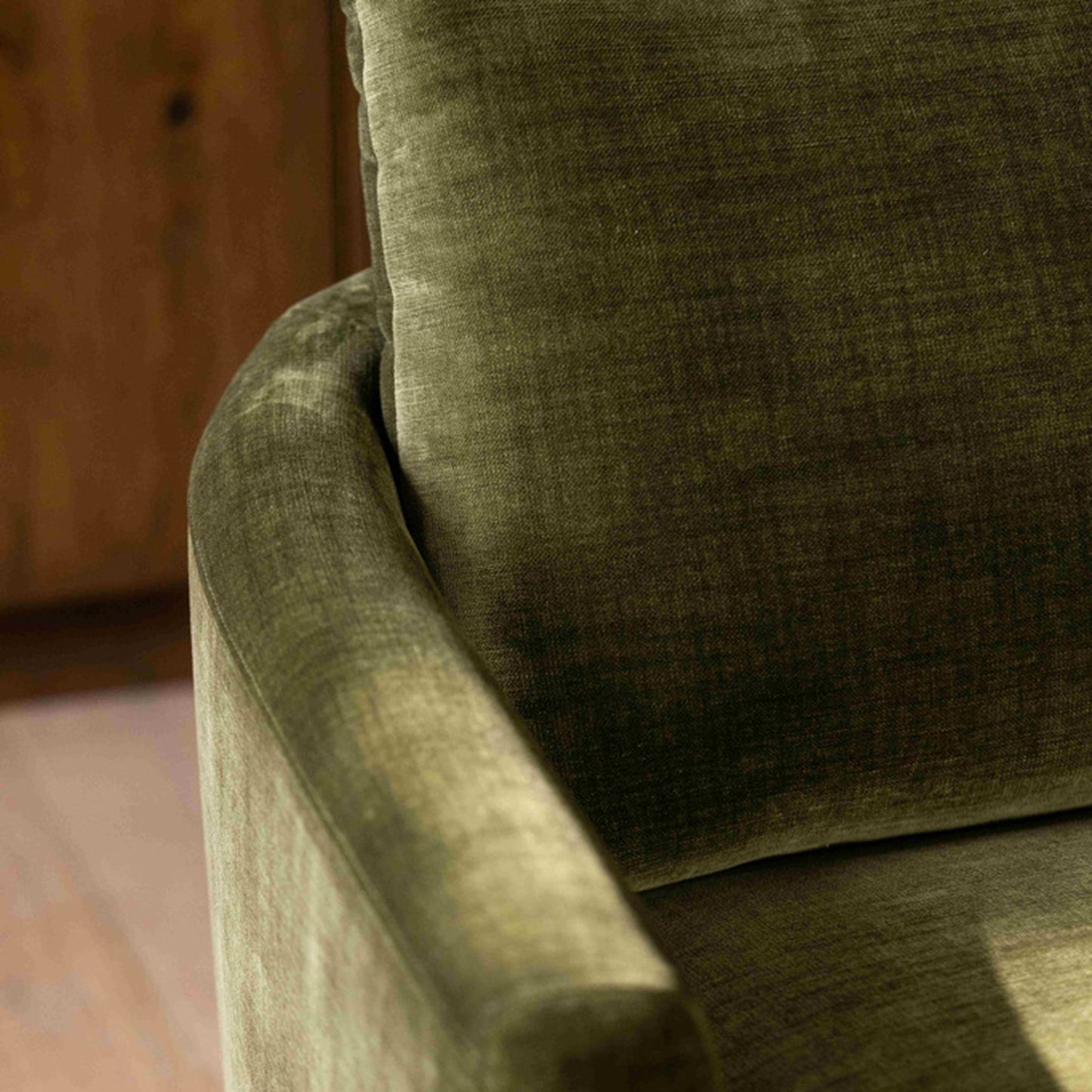 Close-up of a textured olive green upholstered armchair with a matching back cushion, showing the fabric weave and rounded arm detail in warm indoor lighting.