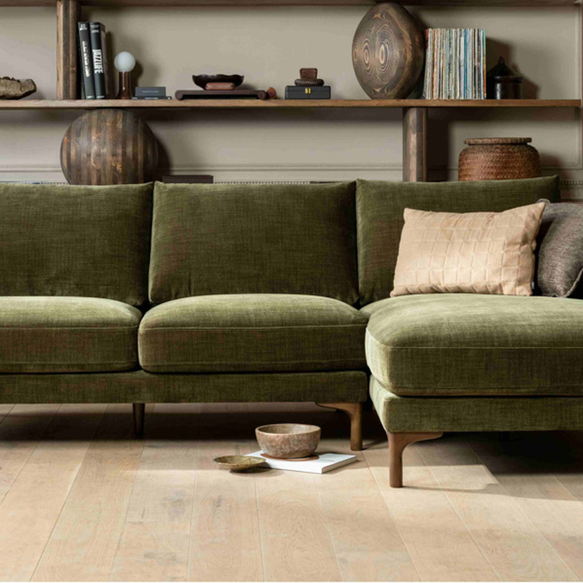 An olive green corduroy sectional sofa with a right-facing chaise lounge, styled in a modern living room with a beige textured pillow, wooden floor, and a wooden bookshelf in the background.