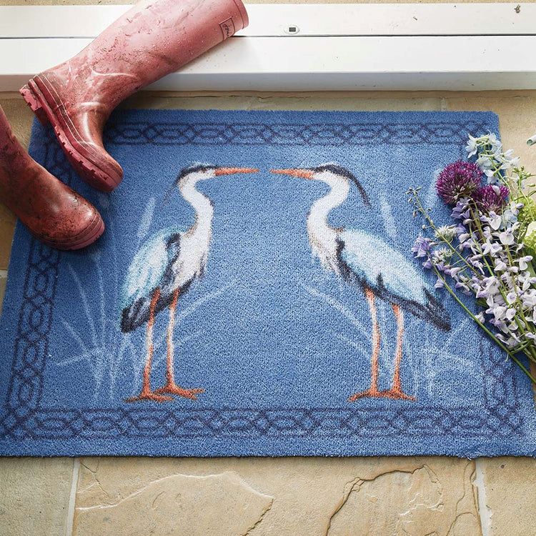 Hug Rug mat at a doorway entrance with two Herons n a blue background with some fresh flowers on one side and brown wellington boots on the other side