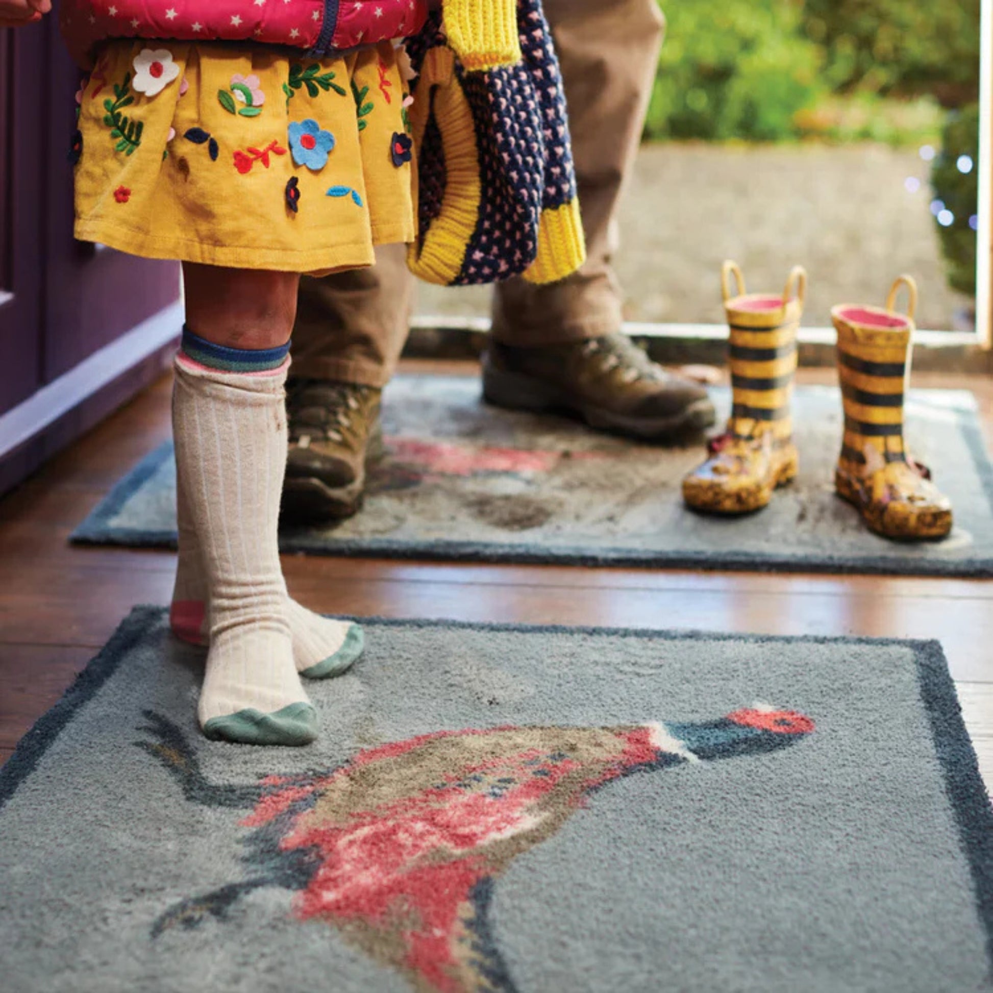 Children's legs in socks and shoes on a rug with a colorful design, next to a pair of rain boots.