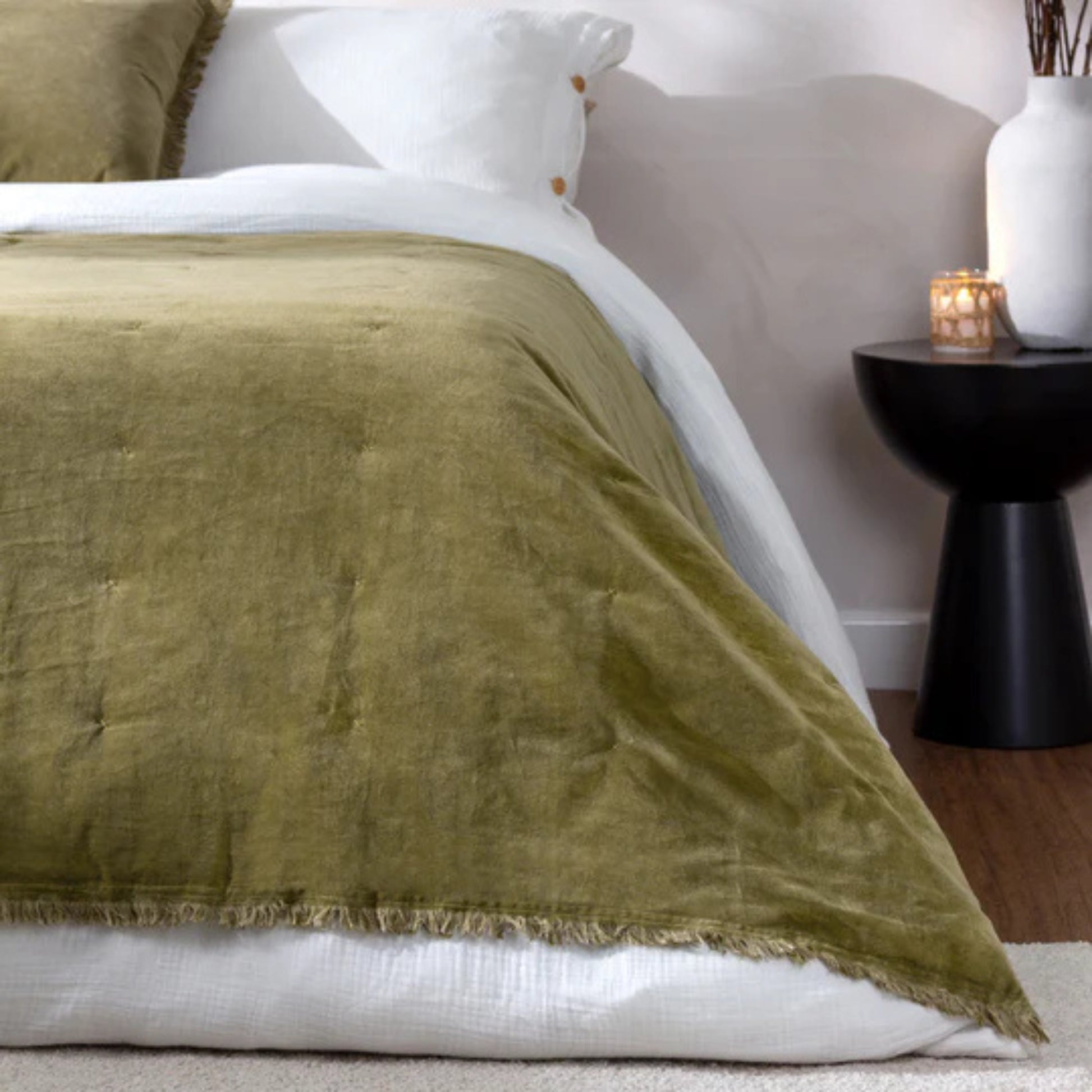 Green bedspread accross a bed dressed in white linen with a dark wood bedside table with a book and two vases sitting on top.