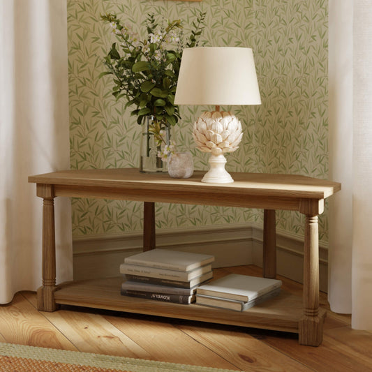 Oak corner tv unit set in the corner of a room with books on the bottom shelf and a lamp and greenery in a glass vase on the top shelf 
