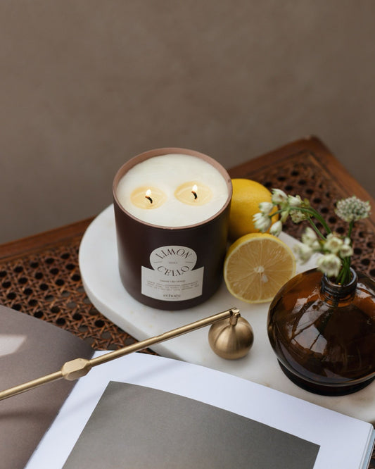 A two-wick candle with a limoncello scent, placed in a brown jar, accompanied by a lemon and some flowers on a table.