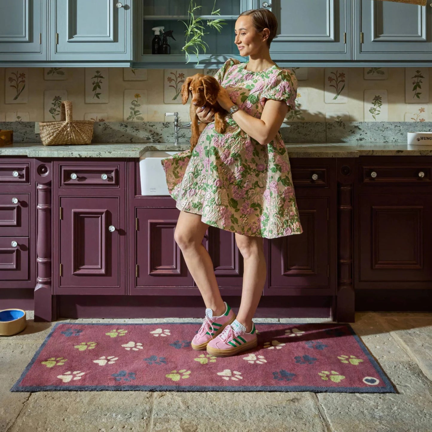 Woman in a floral dress holding a dog in a kitchen with a burgandy rug with a paw print pattern.