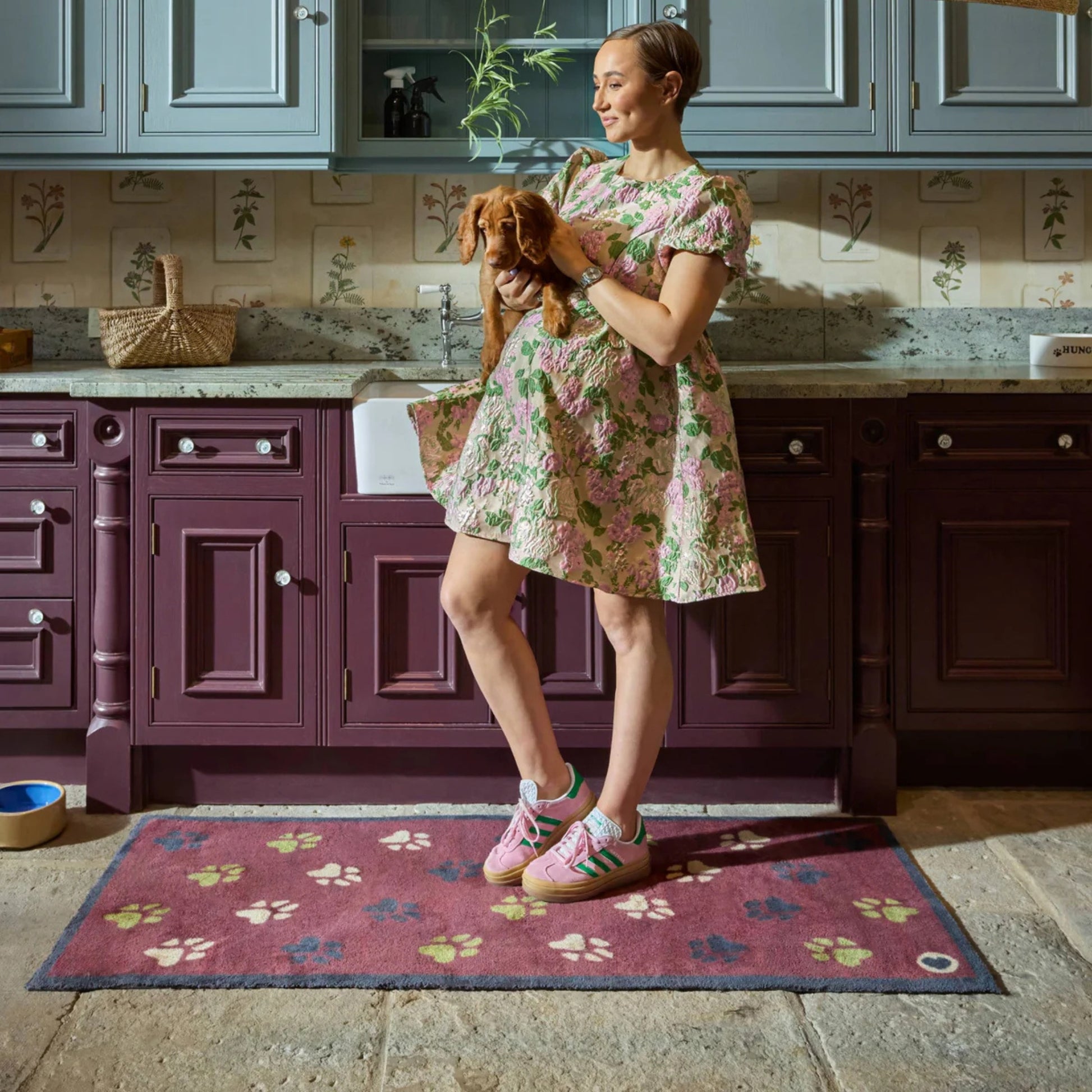 Woman in a floral dress holding a dog in a kitchen with a burgandy rug with a paw print pattern.