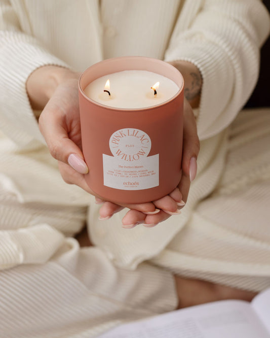 A person holding a pink and lilac colored candle with two wicks, in a jar.