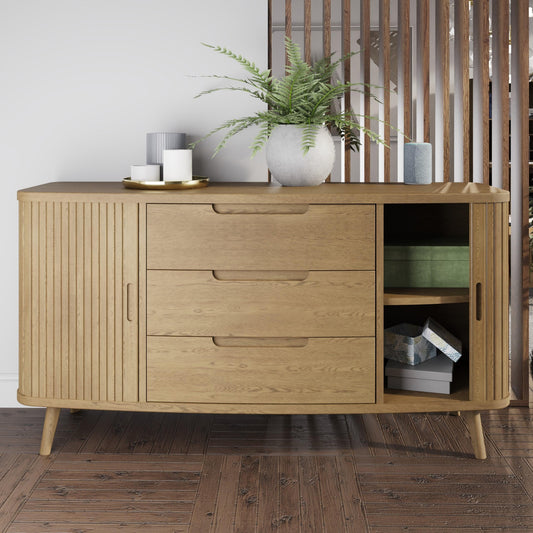 Wood sideboard with palnt pot and candles on top, 3 drawers and one curved sliding door open set against a white wall and slatted wood room divider 