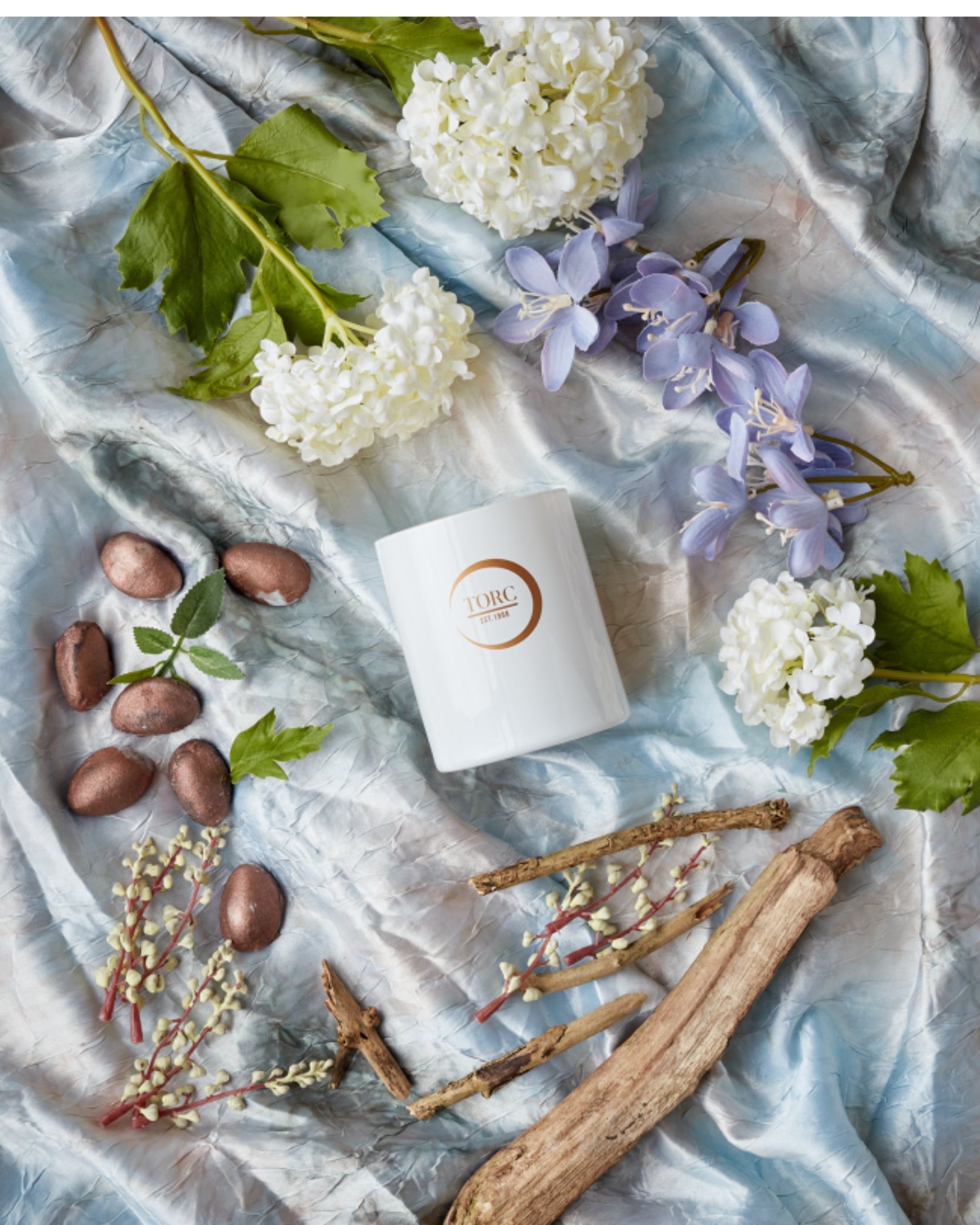 White candle with gold logo surrounded by flowers, leaves, and sticks on a light fabric background