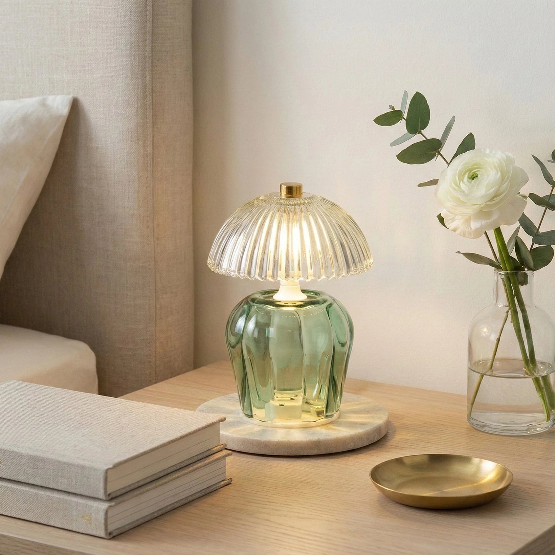 Green glass table lamp on a wooden surface with books and a vase of flowers.