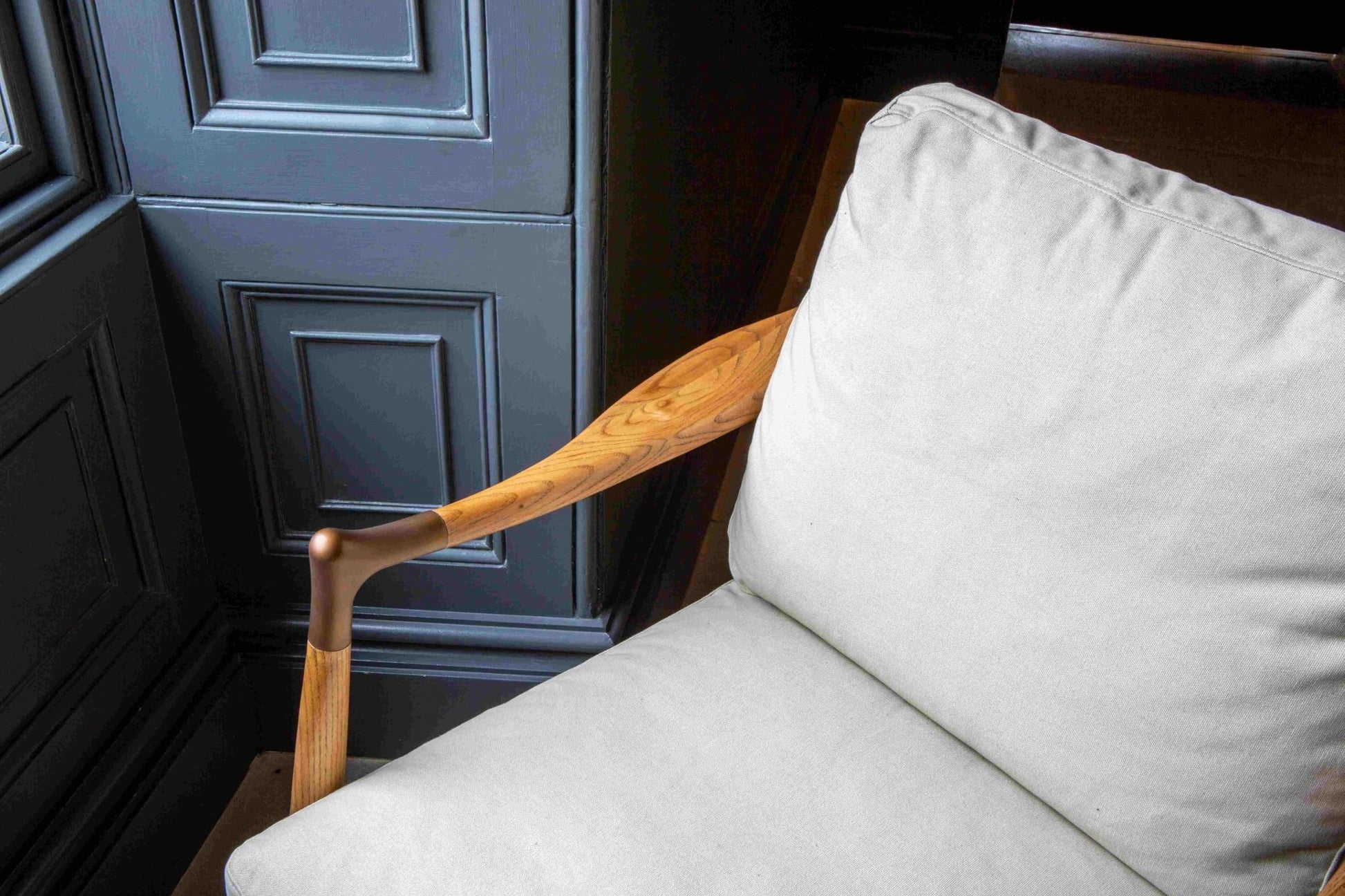 Detail close up of An elegant Archie Accent Chair with a solid ash timber construction and an upholstered ivory canvas seat and back cushion facing a window on a tiled floor with dark navy painted walls. 