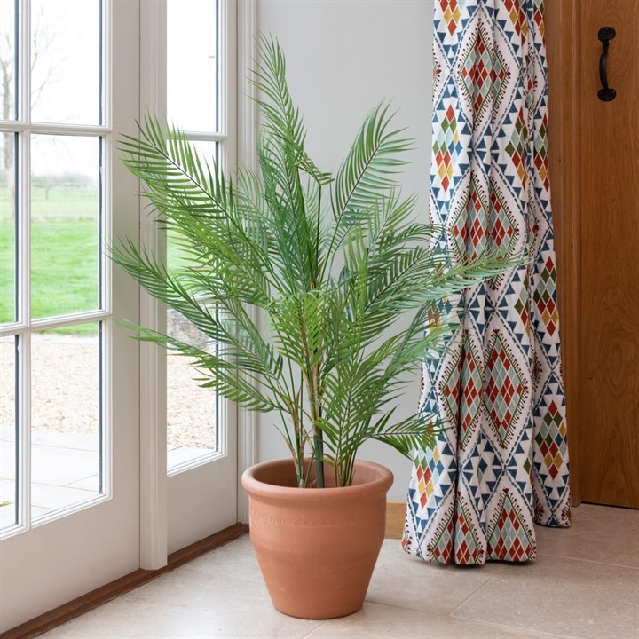 A faux areca palm plant with vibrant green leaves in a terracotta pot sitting on the floor next to window with a colourful floor length curtain. 