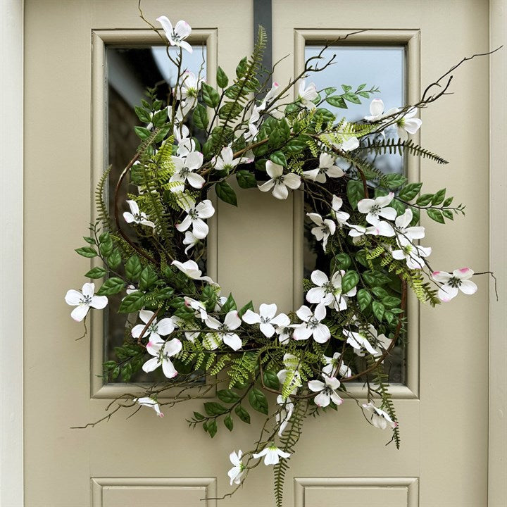 A faux Dogwood & Fern wreath with green leaves and white flowers, hanging on a light green door 