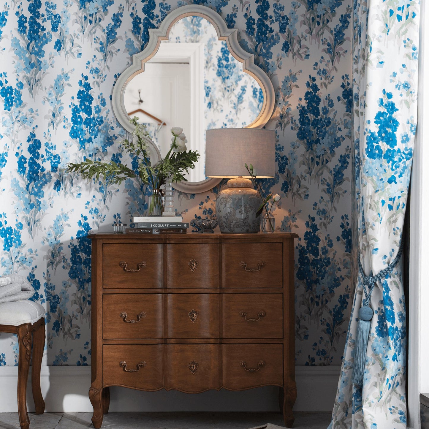 walnut 3 drawer chest styled ina  bedroom featuring blue print wallpaper. Chest is styled with a lamp and vase of flowers with a decorative mirror hanging above it.